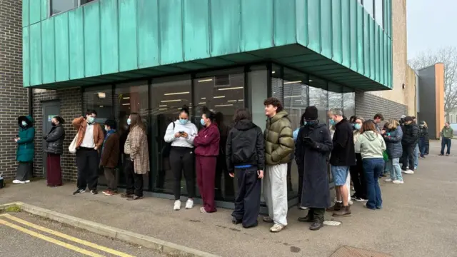 Young people queue around the outside of a modern-looking building.