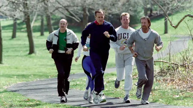 George HW Bush and Chuck Norris and others jog on a paved path in a park, wearing sweats
