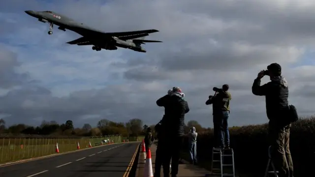 Four people with cameras stand on a roadside photographing a US B1 Bomber