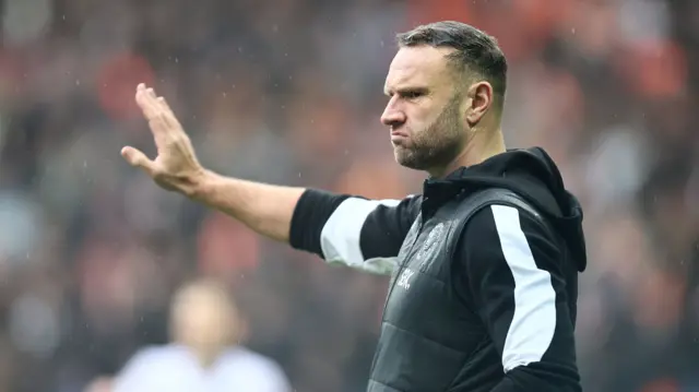 A photo of Ian Evatt holding his arm out while wearing a black long sleeved top with white stripe and a black gillet with the Blackpool club badge on his chest