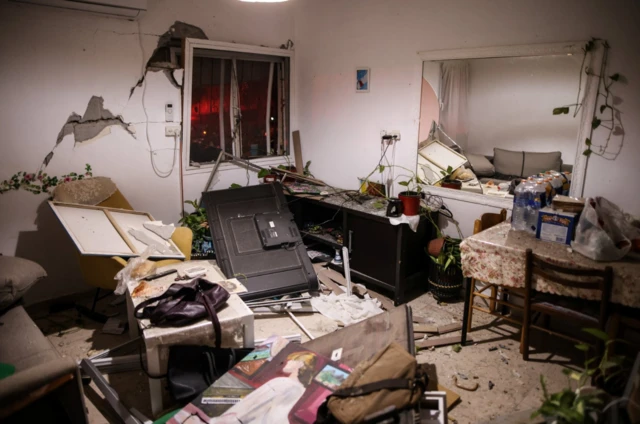 The inside of what appears to be a living room in an apartment bloc in Arad following a missile strike. There's exposed parts of concrete walls without missing chunks of plaster, windows with no glass and furniture overturned on the floor. Debris, including pieces of wood and glass litter the floor