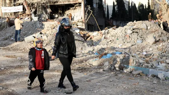 A mother and child walk past a heap of rubble in Tehran. They are both wearing leather jackets and motorbike helmets.