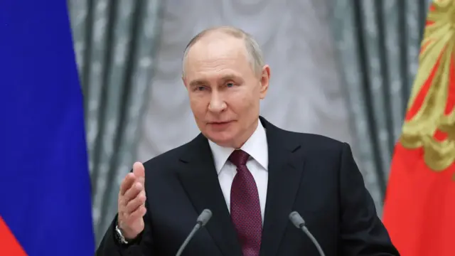 Putin stands in between two flags and speaks into two small microphones.