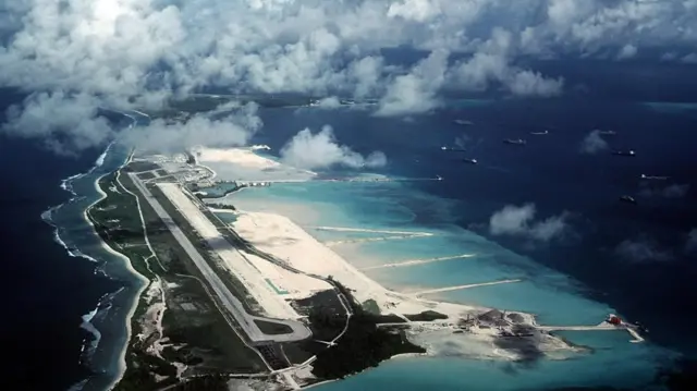 An aerial view shows a runway next to a beach surrounded by clear blue water on a narrow island