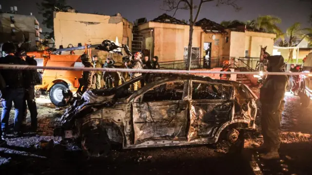 The burnt out shell of a car is shown, with emergency tape in the foreground. workers can be seen inspecting damage and congregate around the car. a house is in the background.