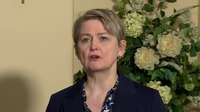 Foreign Secretary Yvette Cooper in black blazer and patterned shirt stands in front of a vase of white hydrangeas and roses during an interview