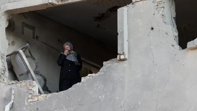 A woman stands inside a destroyed building, covering her mouth and nose with her hijab. she is dressed in black and standing in a window, looking out from the remains of her flat.