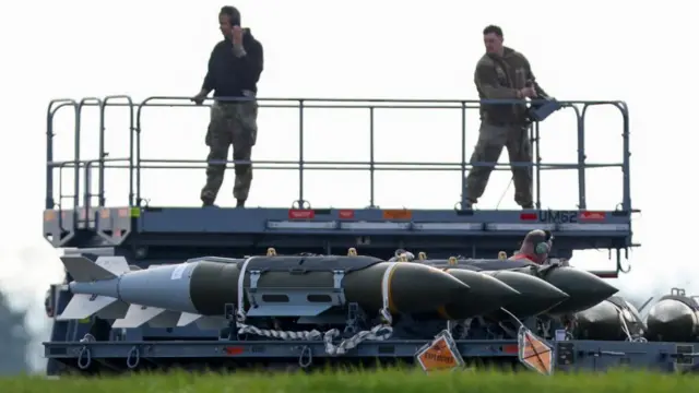 Two members of ground crew stand on a scissor lift next to a row of missiles at RAF Fairford