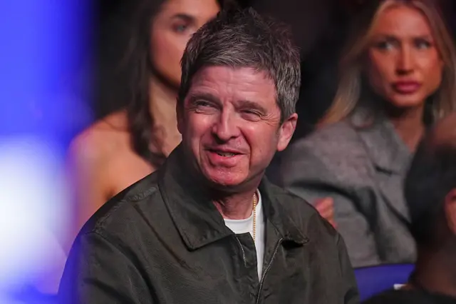 Noel Gallagher smiles and is wearing a grey jacket