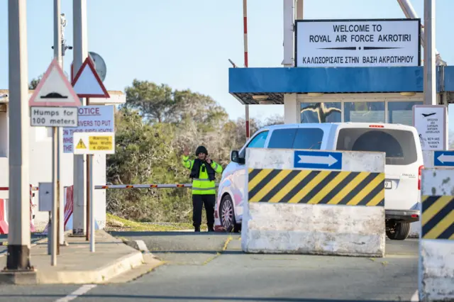White minivan at security check at the entrance of RAF Akrotiri in Cyprus. A guard in a yellow hi-vi jacket stands in front of the van, his right hand raised and speaking on the phone