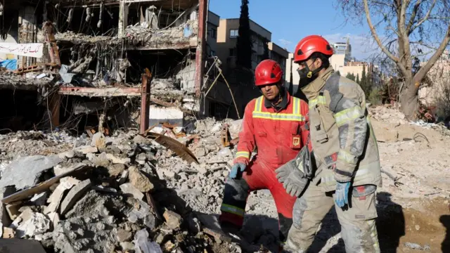 Two emergency workers stand beside a rubble heap. one is dressed in red and one in a beige-brown fireman's uniform. Both wear red hardhats.