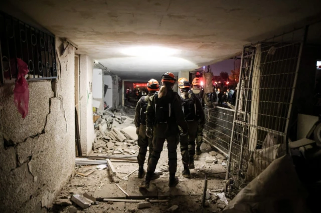 Emergency respondents in heavy duty gear walk along the entrance corridor of an apartment bloc, debris and pieces of wall littering the ground, red lights shining in the distance