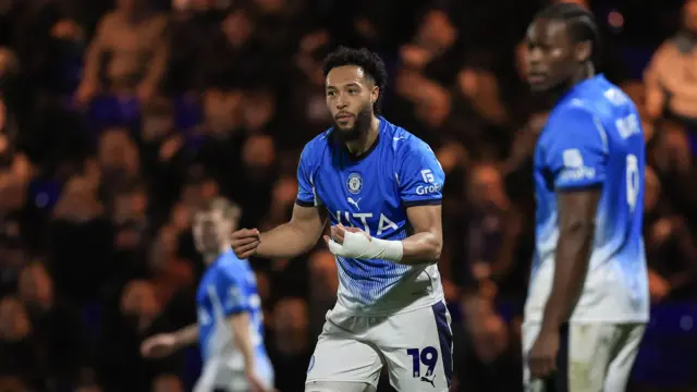 Kyle Wootton reacts with a bandage wrapped around his hand, wearing a blue Stockport County home kit, with his team-mate in the background.