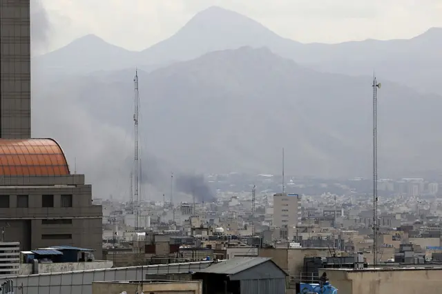Wide shot of Tehran skyline, a cloud of dark smoke billows from a building after a recent strike