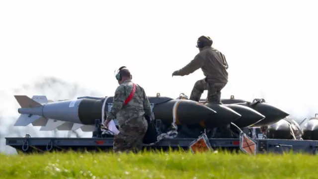 Two ground crew are seen working next to four large missiles near a field of grass. More munitions can be seen in the background