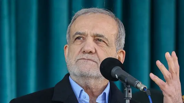 Masoud Pezeshkian mid-shot. He's speaking into a mic, his left hand raised. He's in a black coat and light blue shirt