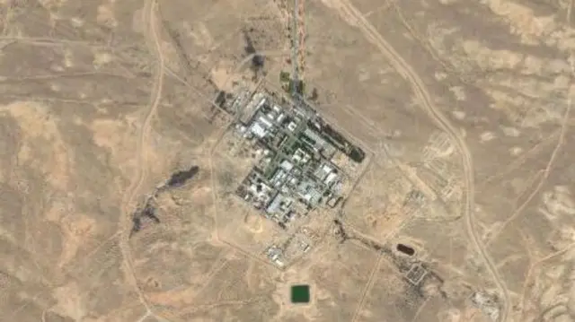 A satellite image shows a small grid of grey buildings in the desert.
