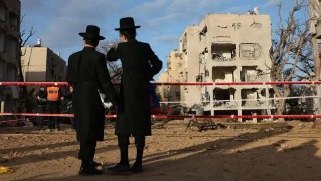 Residents look on at the scene of a direct hit of an Iranian missile in Arad. A building can be seen damaged to the right of the pair