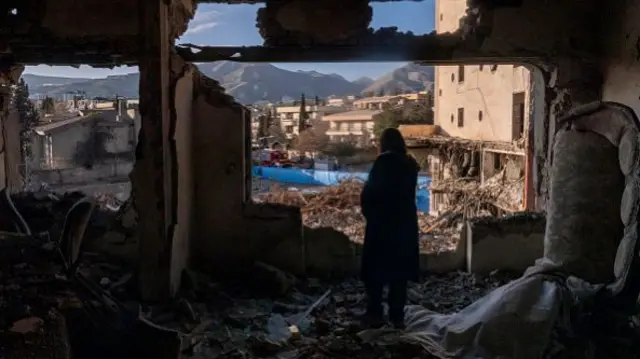 A woman looks out at the hils in the distance from her destroyed apartment in Tehran. A sofa is turned on its side and the wall has been reduced to rubble