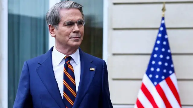 US Treasury Secretary Scott Bessent stands in front of a US flag wearing a blue suit with a bright blue and yellow striped tie