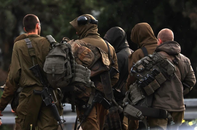 Five Israeli soldiers in military fatigues carrying weapons and equipment walk on a road, their backs to the camera