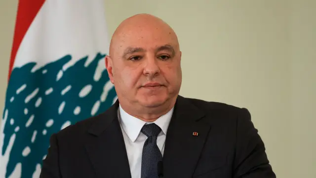 Joseph Aoun in black suit posing in front of a Lebanese flag