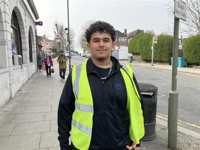 A man wearing a black zip-up hoodie and a reflective vest is shown on a London high street.