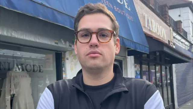 Jack Taub, a member of the leadership team of the Machzike Hadath Synagogue, is photographed in front of shops wearing glasses and a zip-up jacket