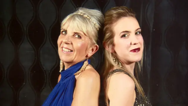 Natalie's mum and Natalie standing back to back. Her mum has blonde hair and blue dress.