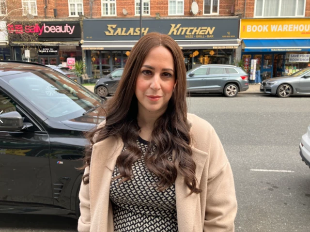 A woman is seen on a high street in front of the road, wearing a black and white dress under a cream overcoat.