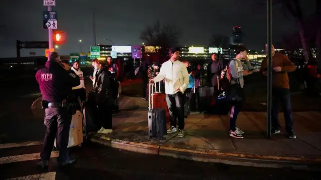 People stand around with suitcases on a pavement while a police officer stands nearby