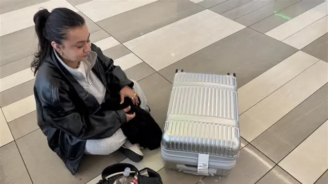 A young woman in a black jacket sits on the ground beside a suitcase.
