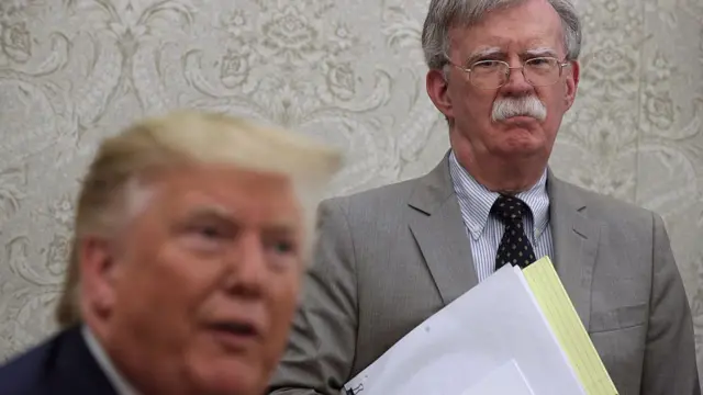 John Bolton standing behind Trump holding a stack of documents.