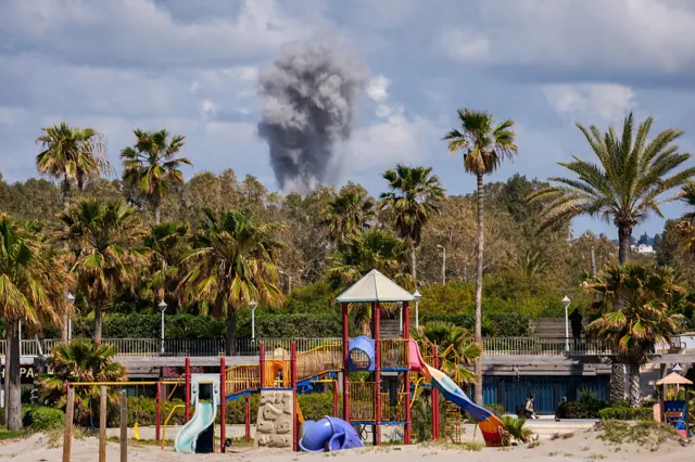 A playground stands empty as smoke rises in the distance from the site of an Israeli airstrike that targeted the outskirts of the southern city of Tyre, with palm trees seen across the foreground and a blue sky around the rising smoke in the background.
