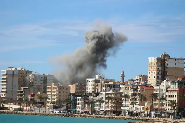 Smoke rises an Israeli airstrike that targeted the eastern outskirts of the southern Lebanese coastal city of Tyre, with a blue sea and sky visible around the buildings.
