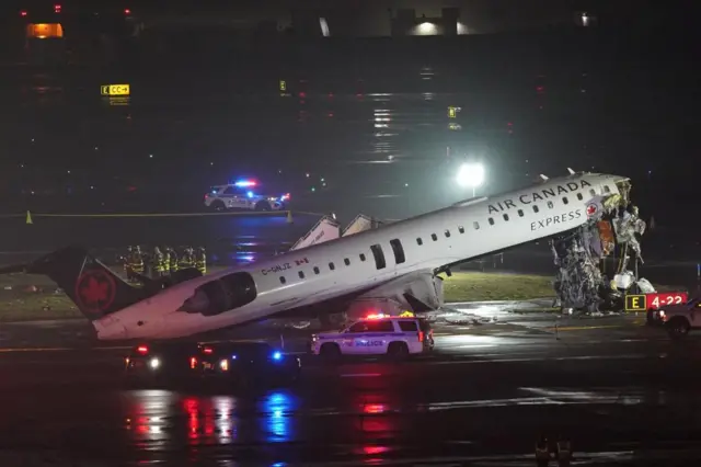 An Air Canada plane with its cockpit missing tilts backward on its tail with emergency vehicles nearby