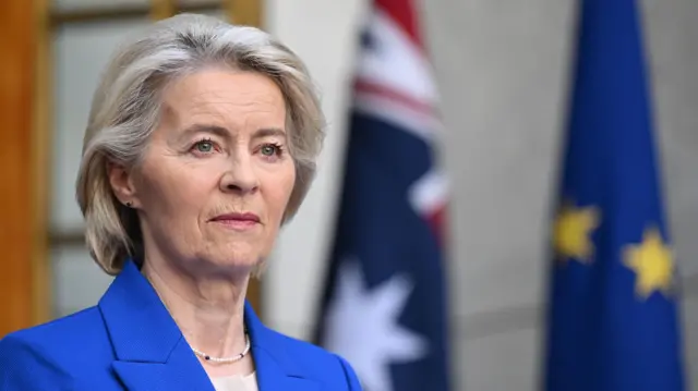 Ursula von der Leyen in a bright blue blazer standing in front of an Australian flag and European Union flag, which are blurred in the background.