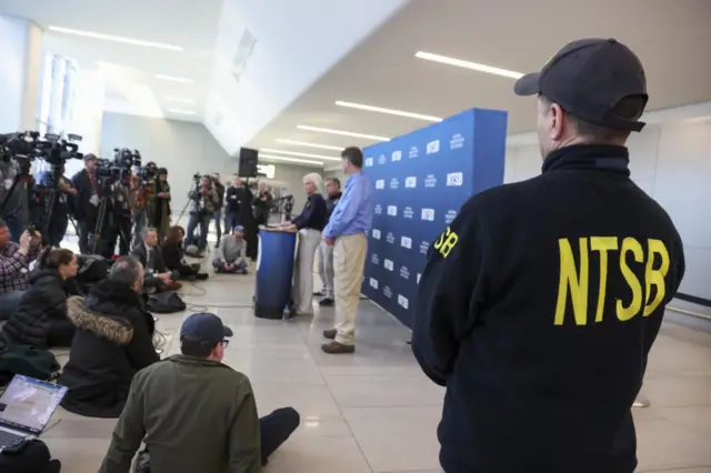 A man in a cap and an NTSB jacket watches as officials brief the press
