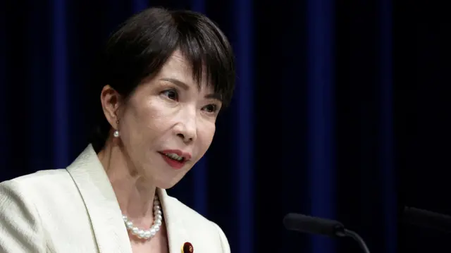 Japan’s Prime Minister Sanae Takaichi speaking into a microphone while wearing a white blazer and pearl necklace.