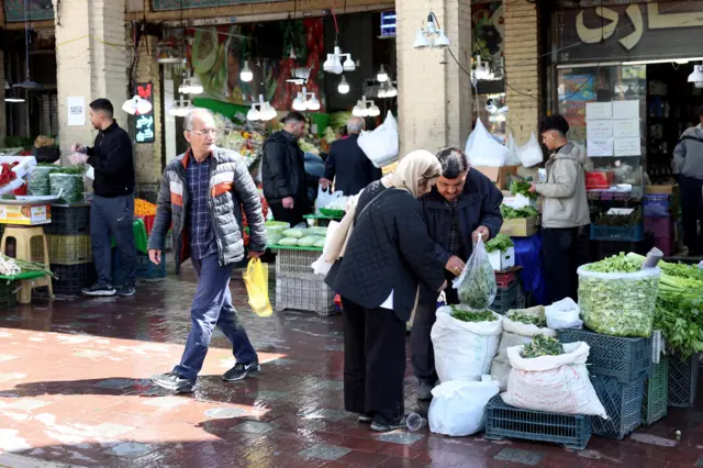 Men and women shops and speak with vendors at Tajrish Bazaar, including fresh green produce and peppers.