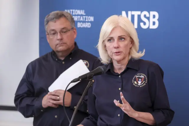 A man and a woman stand in front of a blue background with NTSB written across it