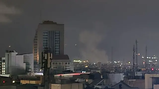Grey smoke rises against a dark sky in Tehran, Iran, with multi-storey buildings visible in the foreground.