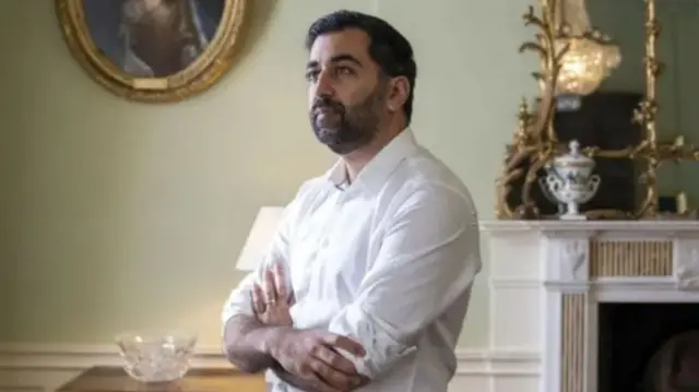 Humza Yousaf, arms folded, stands thoughtfully in Bute House