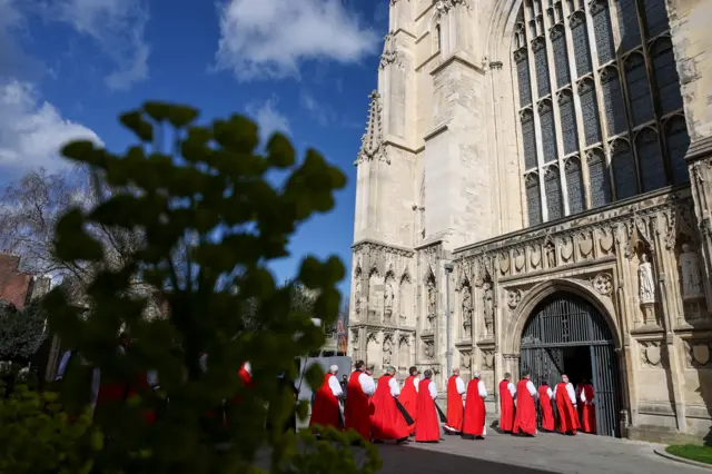 Red robed religious crowds enter from exterior of cathedral