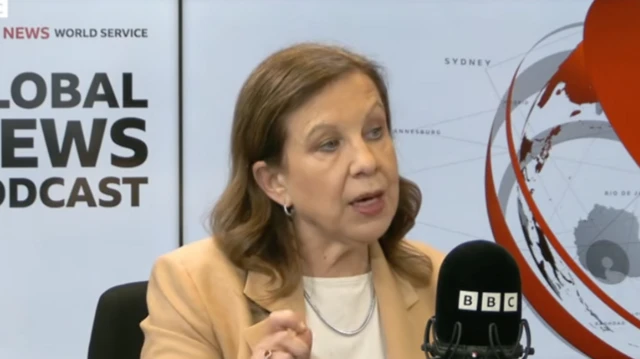 A woman in a white top and beige jacket is seen talking into a black microphone labeled "BBC".