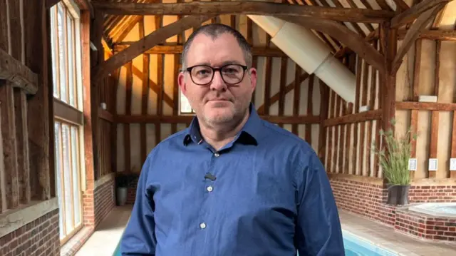 Spa owner Daniel Pilley looks to camera while standing in front of an indoor pool and hot tub