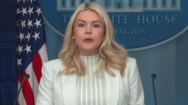 White House Press Secretary Karoline Leavitt wears a white jacket and blouse and speaks with the US flag behind her.