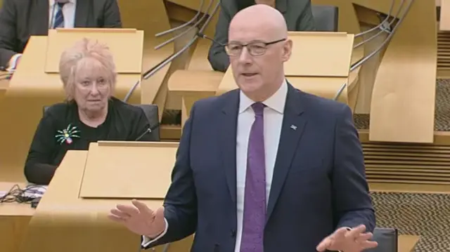 Christine Graham sits behind a standing First Minister John Swinney in her regular seat in Holyrood