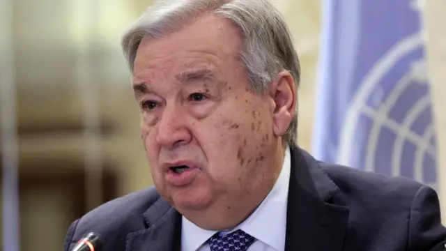 Antonio Guterres speaks into a microphone in front of a UN flag