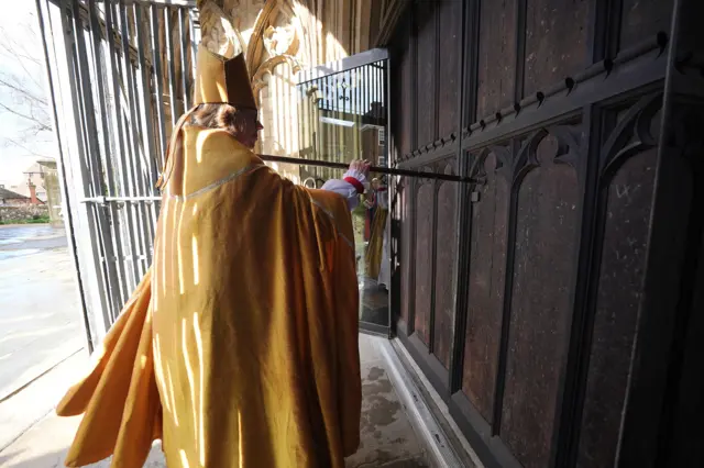Sarah M in yellow robes knocks on cathedral door with staff